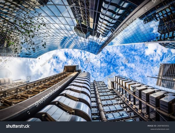 An up shot of buildings up towards the sky during slightly cloudy weather. The buildings are extremely reflective and reflect the clouds from the sky